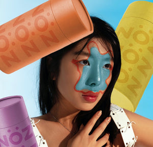 Headshot of a woman wearing blue and orange Nöz SPF on her face with purple, yellow, and orange Nöz sunscreen sticks in the background.